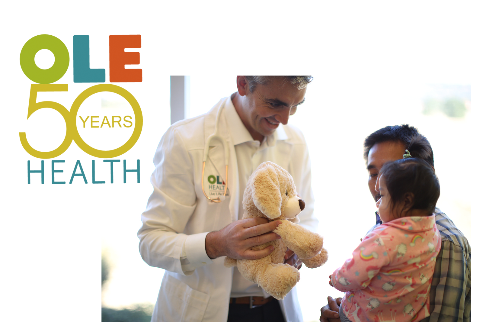 OLE Health 50 Years - a doctor handing a stuffed dog to a child that is being held by her dad.
